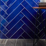 4x16 Gioia Navy colored tile set in a herringbone pattern on a wall close up abutting a wood-look floor.